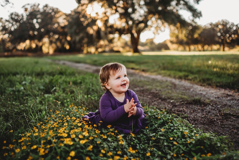 Family Photography, a baby kneels and smiles while sitting among wildflowers in the grass