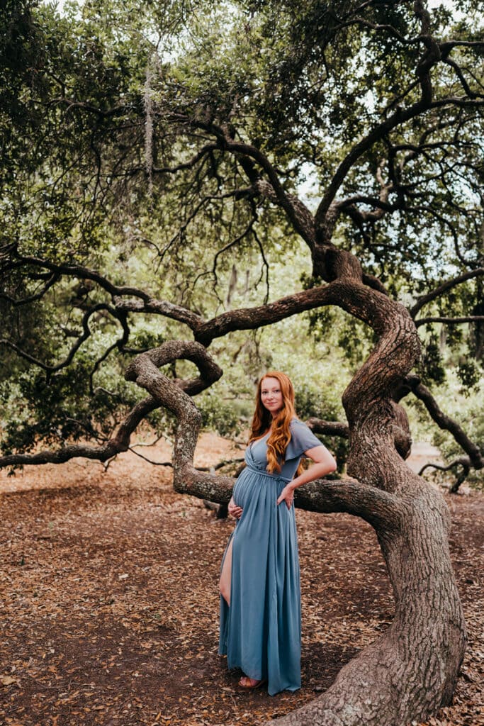 Maternity Photographer, an expectant mother stands underneath a tree with hand on belly and happy