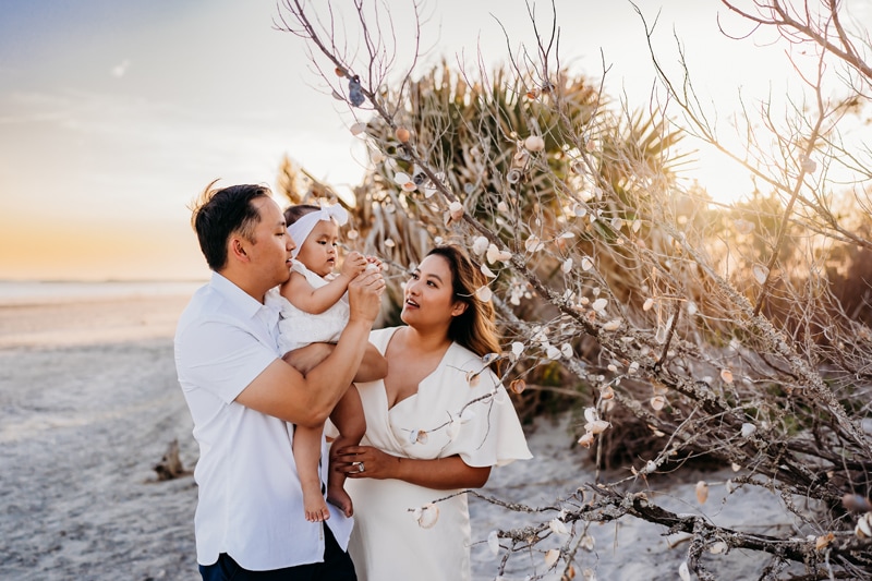 Family Photographer, at the beach, dad holds baby as mom looks on at golden hour